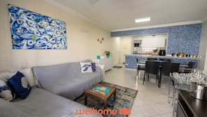 A modern living room with a sofa, coffee table, and a painting. Adjacent is a kitchen with a tiled backsplash and a dining area with chairs. - Apto em Riviera de São Lourenço a 300m da Praia (Bertioga)