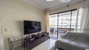 A living room with a flat-screen TV, a glass coffee table, a sofa, and a balcony with a view of buildings. - Apto em Riviera de São Lourenço a 300m da Praia (Bertioga)