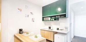 Una cocina compacta con microondas, lavamanos y un refrigerador pequeño. Hay una mesa de madera con una cafetera y un jarrón. Los armarios son verdes y la encimera es blanca.