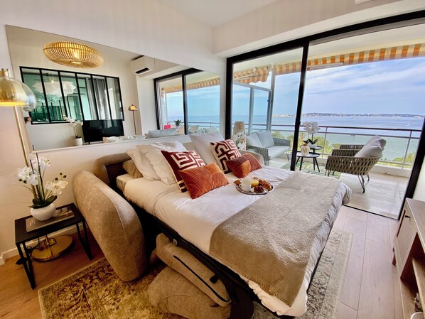 Quarto moderno com cama grande, varanda com vista para o mar e área de jantar com mesa e cadeiras.