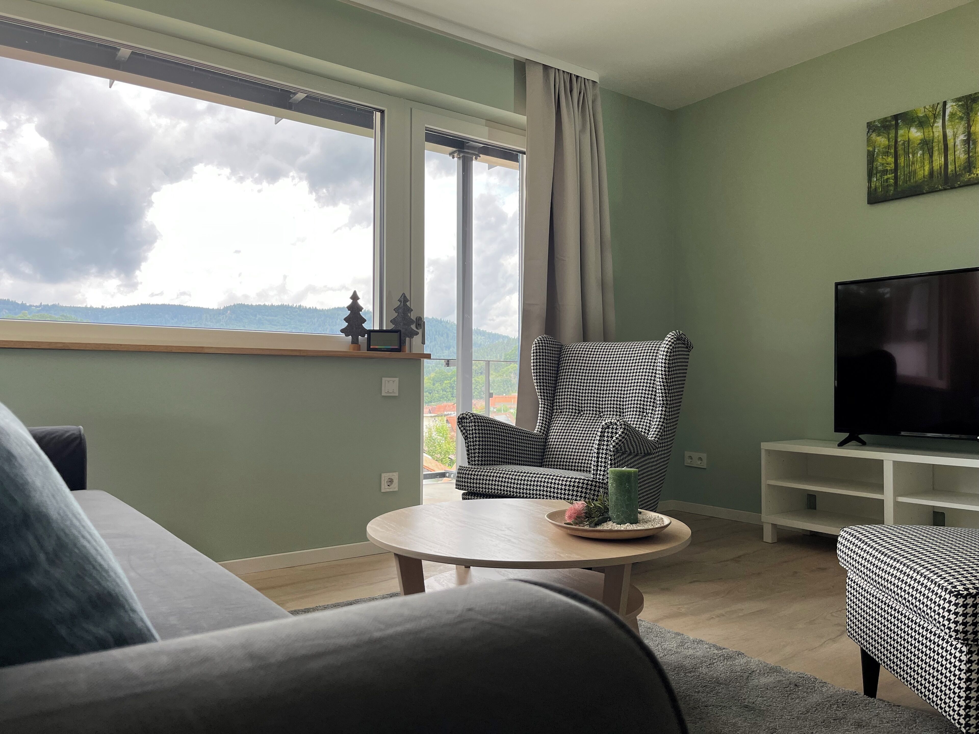Ein Wohnzimmer mit einer Couch, einem hundszahn-gemusterten Sessel, einem runden Couchtisch mit einem dekorativen Gegenstand, einem Fernseher auf einem Schrank und einem großen Fenster mit Blick auf Berge und Wolken.