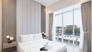 A hotel room with a large bed, a nightstand with a lamp, a window with curtains, and a view of buildings outside. - StayShort - Comfortable 1BR Apartment in JVT with Gym & Pool Sleeps 2 (JVT)
