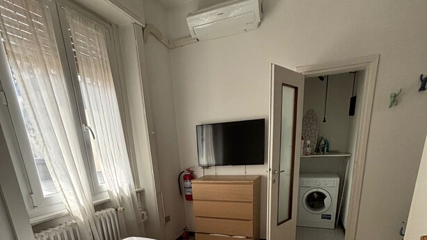 A bedroom with a bed, a TV, a dresser, a washing machine, and a bathroom visible through an open door. - Cosy Studio Quick Metro Ride to Centre (Milan)