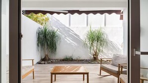 Een modern terras met een houten tafel en twee rieten banken, omringd door planten in potten en een witte muur.