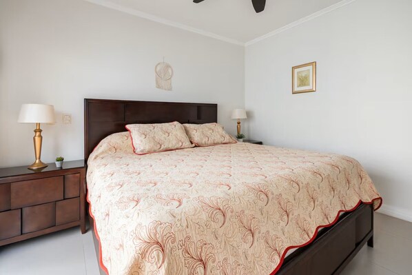 Un dormitorio con una cama grande, cabecera de madera, mesitas de noche con lámparas, una planta pequeña y un cuadro en la pared.