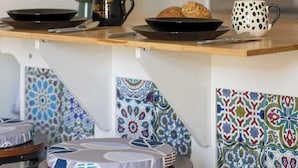 Una encimera de cocina con una cafetera, una taza y un plato de galletas. Hay dos taburetes con cojines estampados. La pared tiene diseños de azulejos coloridos.