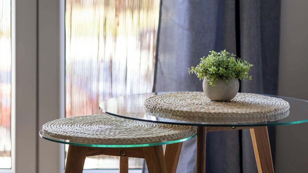 Una mesa con cubierta de vidrio, un mantel tejido, una planta en maceta y patas de madera.