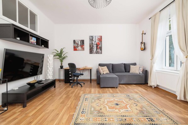 A modern living room with a flat-screen TV, a grey sofa, a desk with a chair, and a decorative rug.