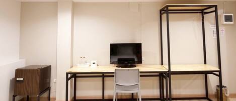 Una habitación compacta con un escritorio de madera, un monitor negro, una silla blanca y un mueble pequeño.