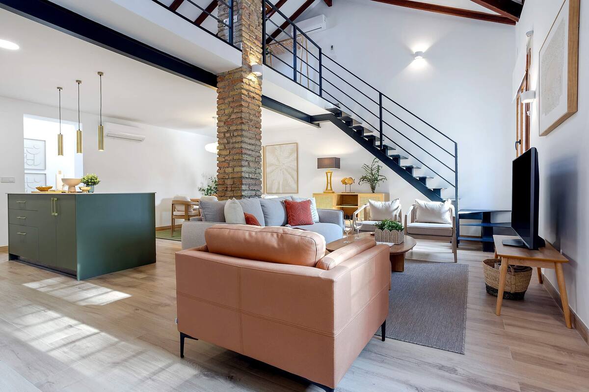 Ein geräumiger Wohnbereich mit hoher Decke, einer Treppe, einem grünen Schrank, einem beigen Sofa und einem Fernseher auf einem Holzboden.