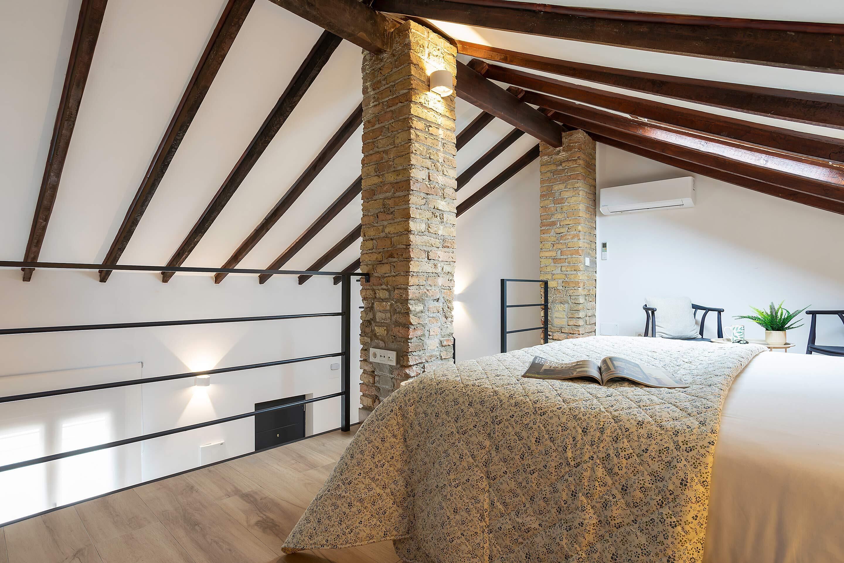 Un dormitorio con techo inclinado, vigas de madera a la vista, un pilar de ladrillo, una cama con colcha estampada y una mesita de noche con una planta, además de una ventana.