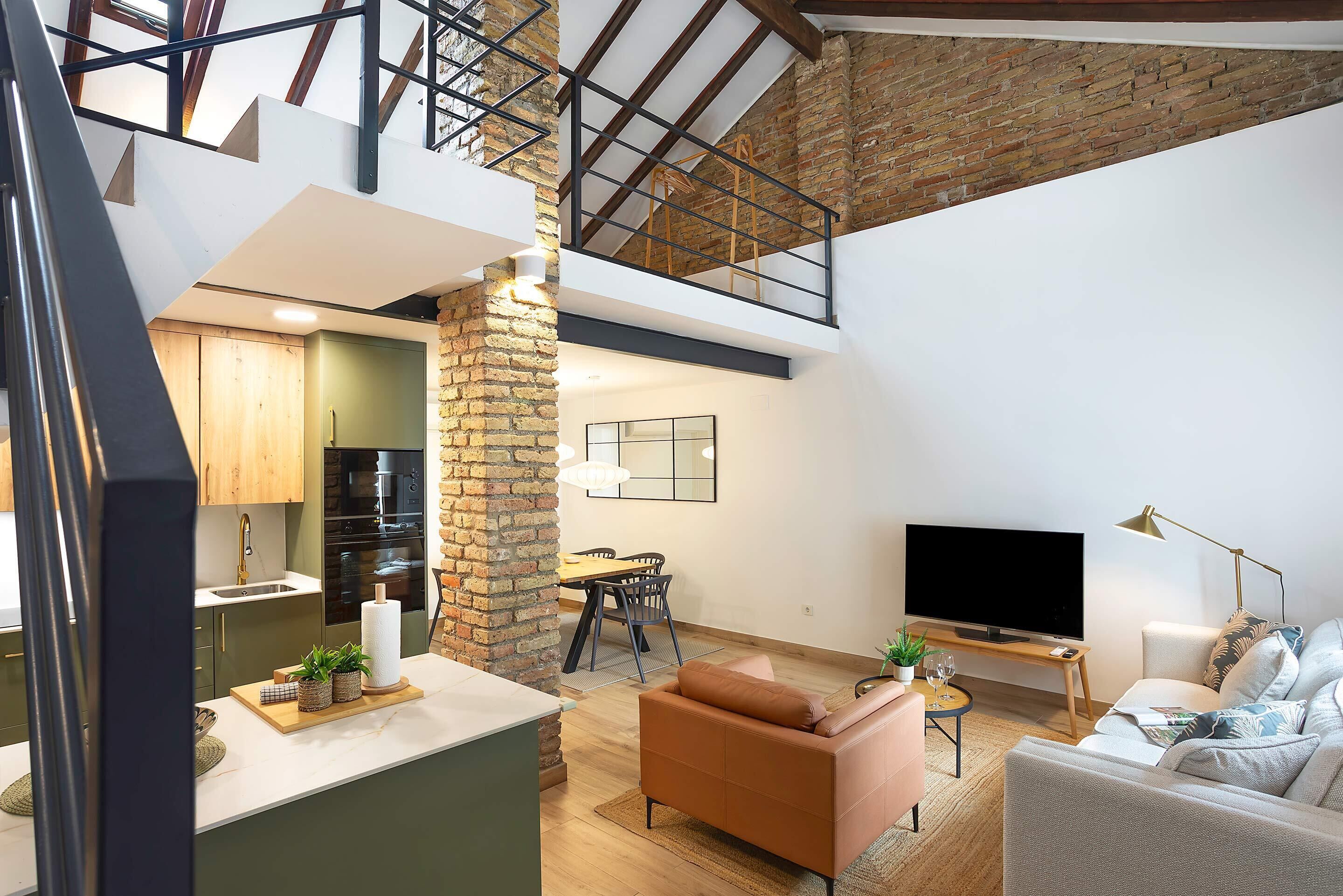 Ein modernes Loft mit Küche, Essbereich und Wohnzimmer, das unverputzte Backsteinwände, Holzbalken und einen großen Fernseher zeigt.