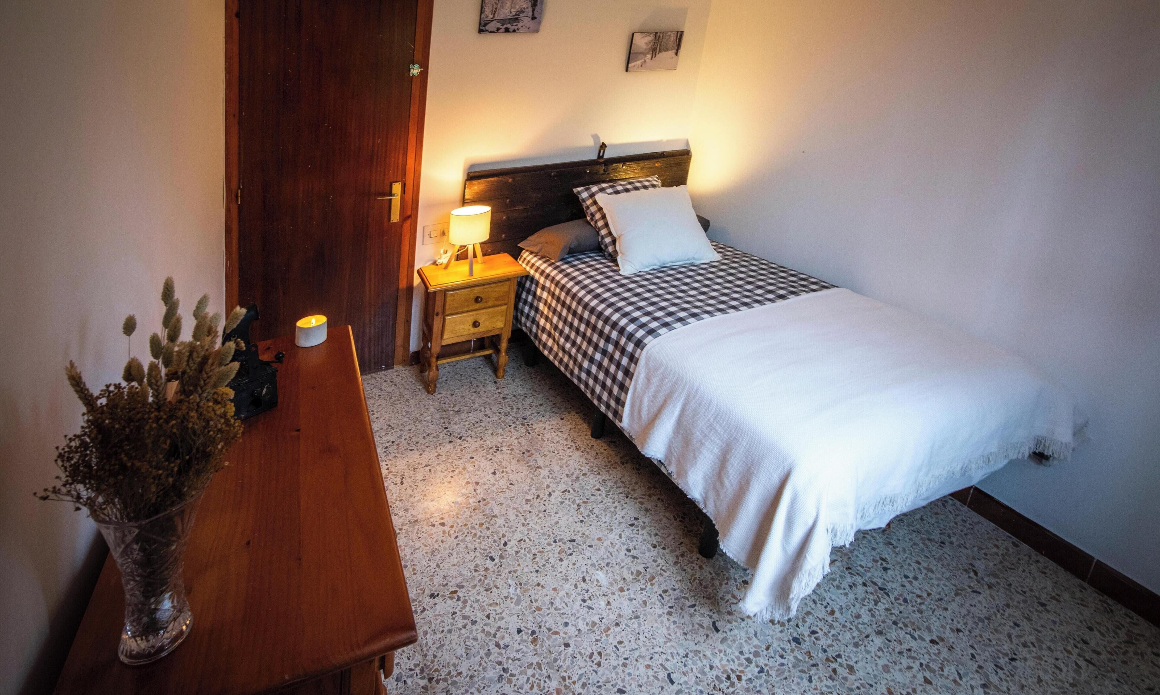 Ein Schlafzimmer mit Bett, Nachttisch, Lampe und einer Vase mit getrockneten Blumen.