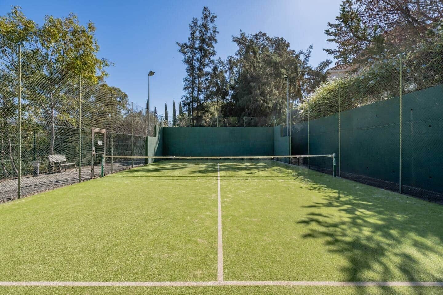 A tennis court with a green surface, surrounded by a chain-link fence and trees.