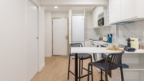 A compact kitchen with white cabinets, a marble backsplash, a white refrigerator, and a small dining area with two stools. - Waou Malasaña VII - Short Term Rental (Madrid)