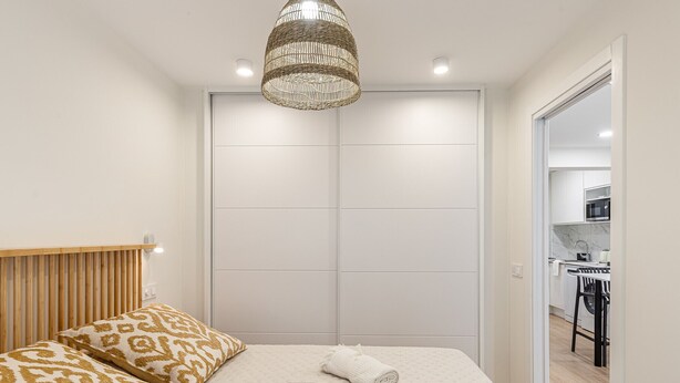 A bedroom with a bed, a bedside table, a hanging light fixture, and a view into a kitchen area. - Waou Malasaña VII - Short Term Rental (Madrid)