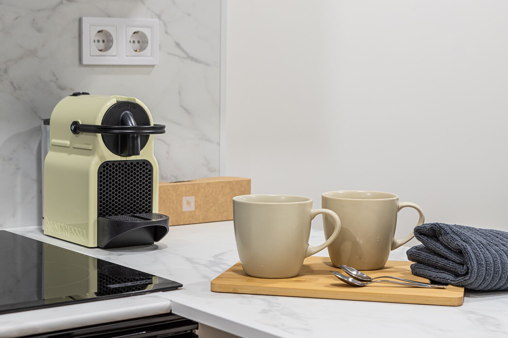 Een aanrecht met een koffieapparaat, twee mokken, een doos, een tablet en een handdoek.