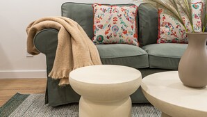 A living room with a green sofa, floral cushions, a beige throw blanket, and two round side tables. - Waou Malasaña VII - Short Term Rental (Madrid)