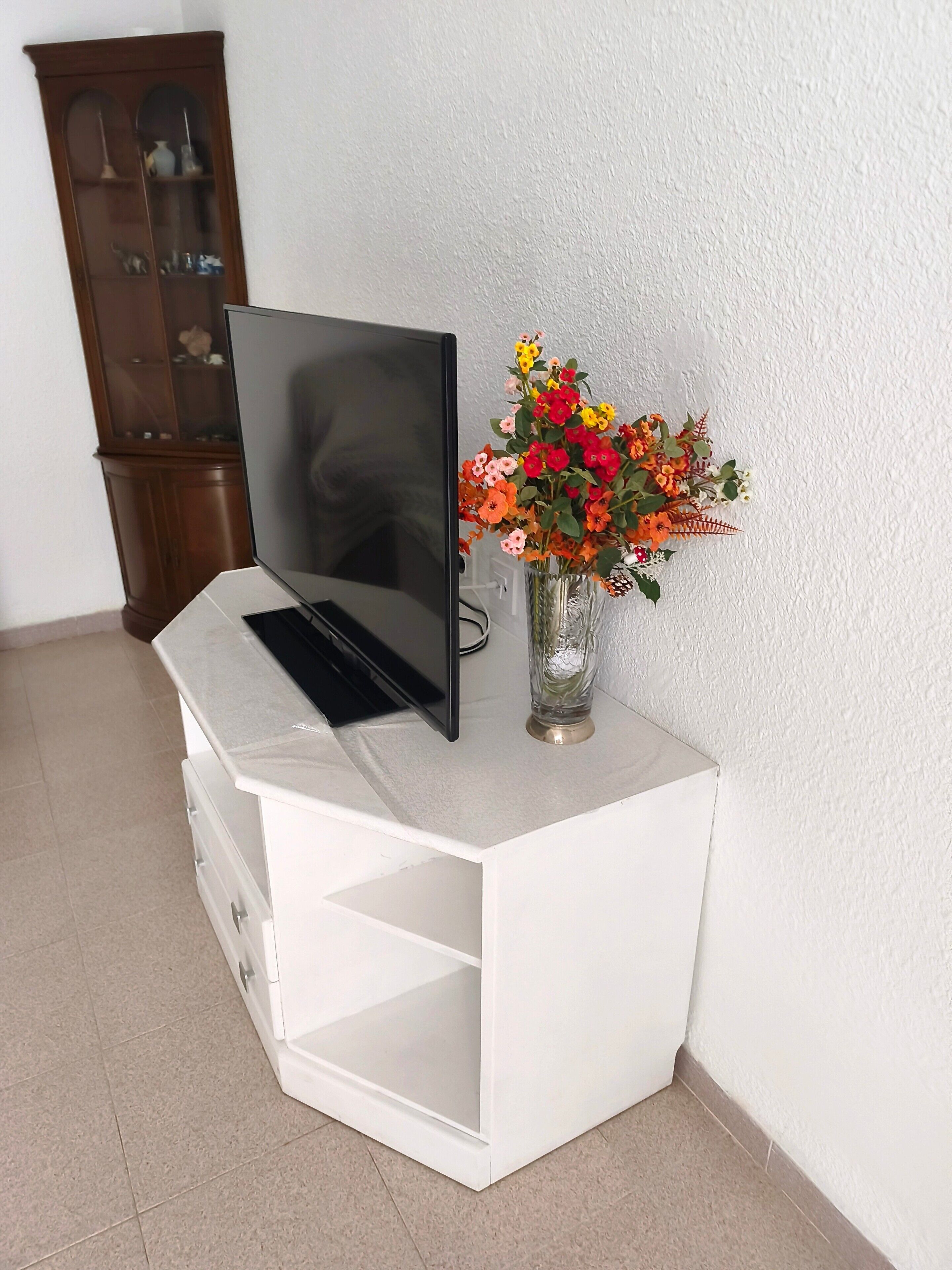 Un mueble blanco de entretenimiento con un televisor de pantalla plana, un jarrón con flores y un armario de madera al fondo.