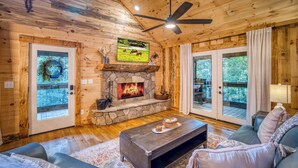 L’intérieur chaleureux d’un chalet avec une cheminée en pierre, des murs en bois, un ventilateur de plafond et une grande fenêtre.
