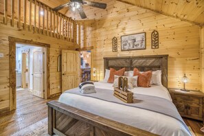 Quarto de cabana de madeira com cama, mesa de cabeceira, ventilador de teto e cozinha visível através de uma porta aberta.