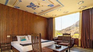A bedroom with a bed, two chairs, a table, and a large window with a view of a mountainous landscape. - Badi Garh Resort (Udaipur)