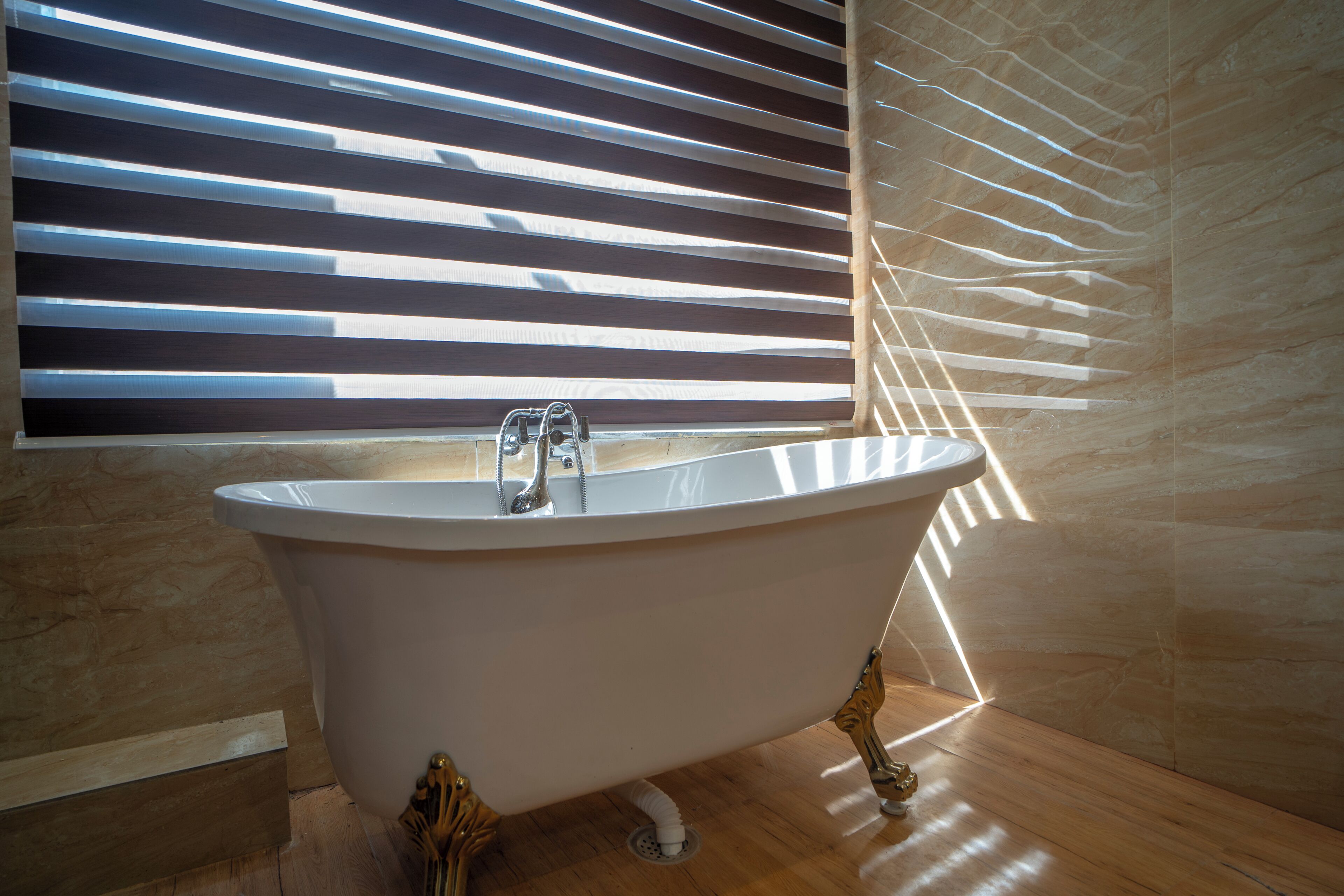 A freestanding bathtub in a bathroom with wooden flooring and a window with horizontal blinds.