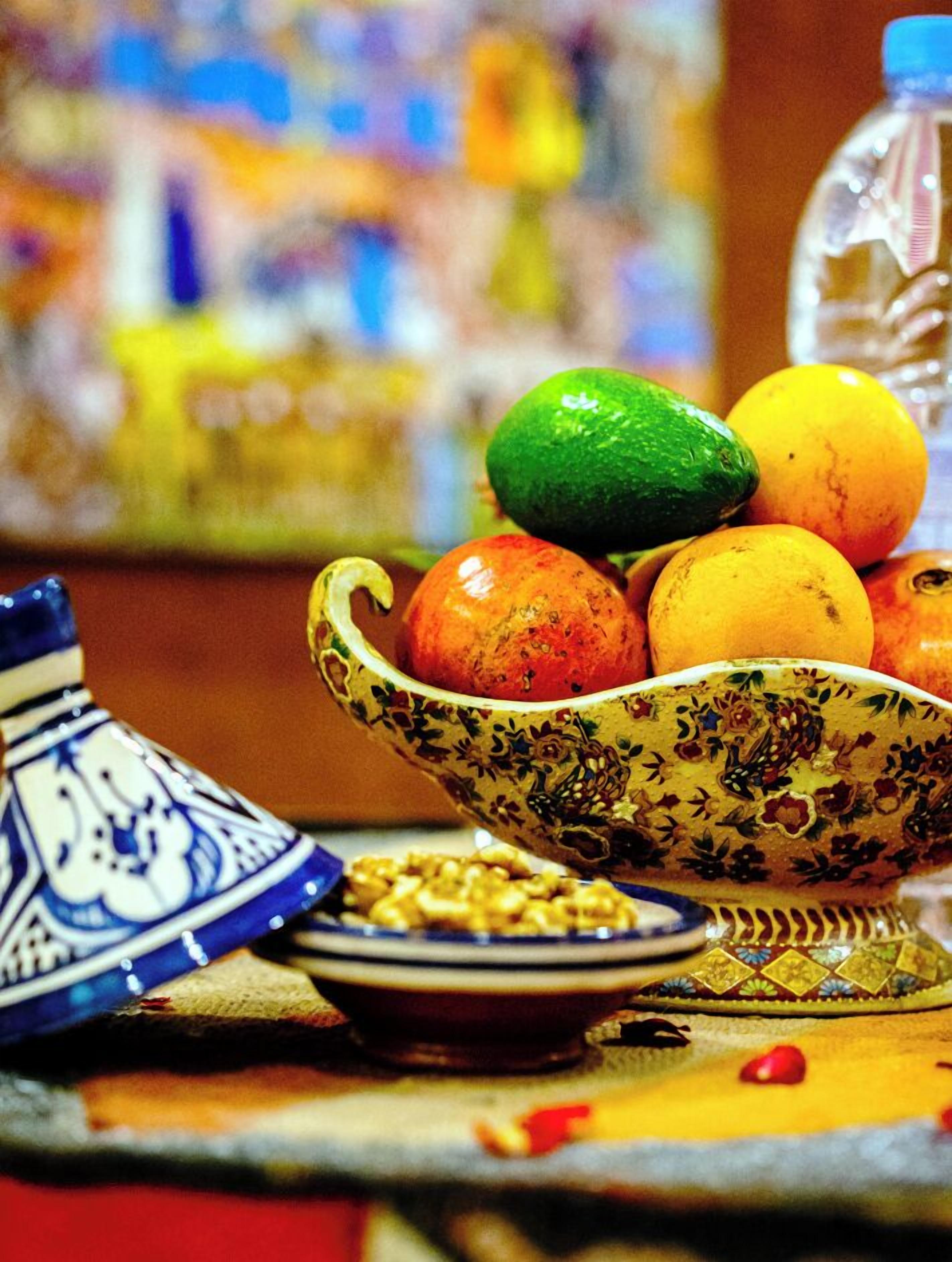 Una mesa con un cuenco decorativo lleno de frutas, un tagine marroquí tradicional y una botella de agua.