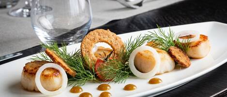 A white plate with seared scallops, a fried item, and garnishes on a dark table.