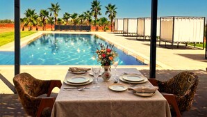 A dining table set for two with a pool view, palm trees, and a clear sky. - Bab Sma - Porte du Ciel (Tameslouht)