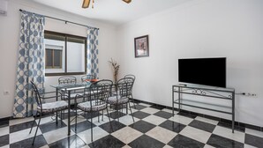 A dining area with a checkered floor, a glass table, metal chairs, a ceiling fan, and a mounted TV. - \"marina house\" conil (Conil de la Frontera)