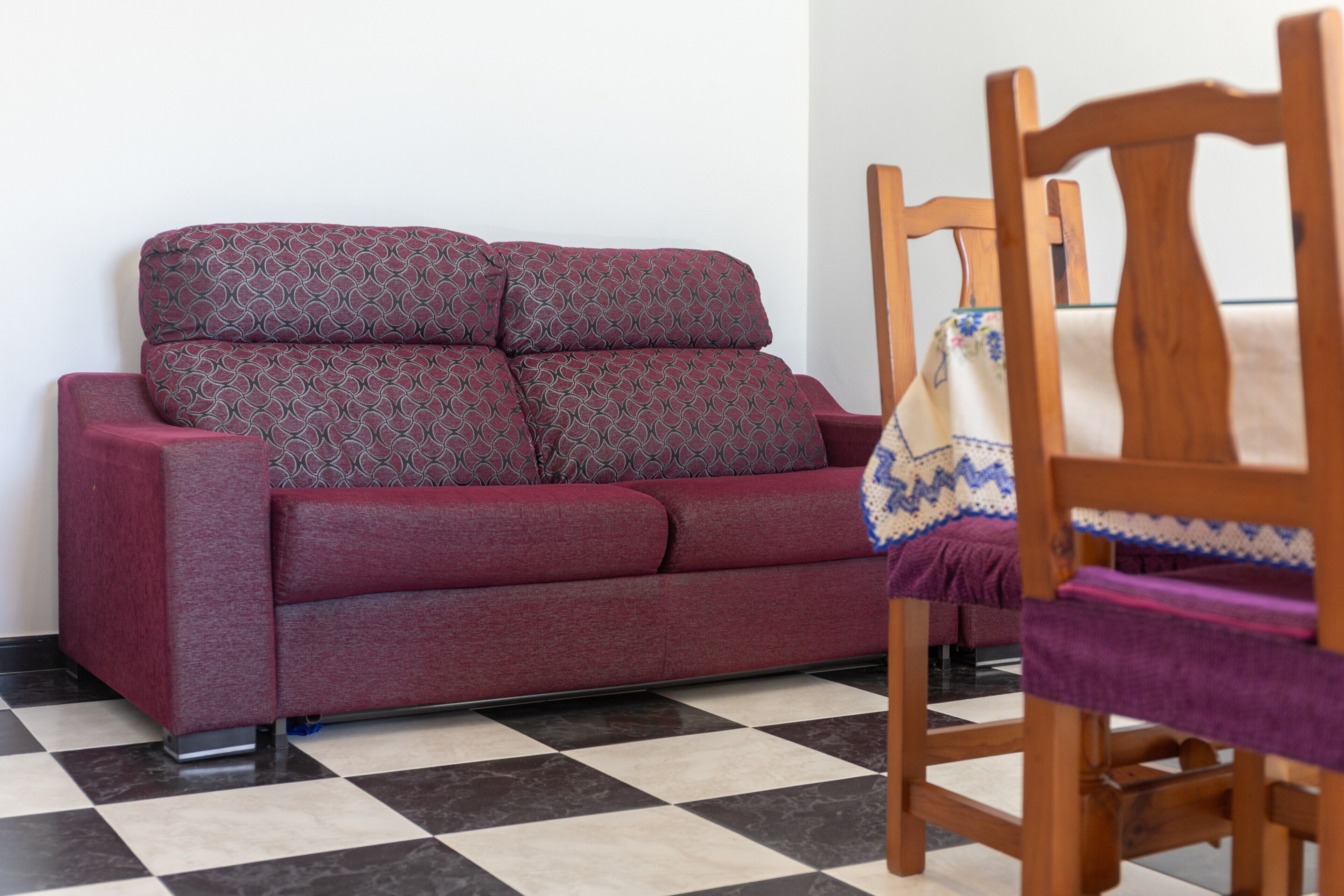Un sofá rojo con cojines estampados, una silla de madera con un cojín morado y un suelo a cuadros.