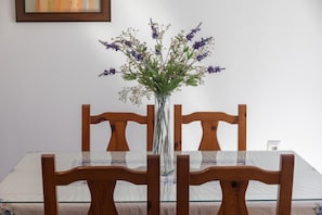 A dining table with a clear glass top, surrounded by wooden chairs, and a vase with purple flowers in the center.