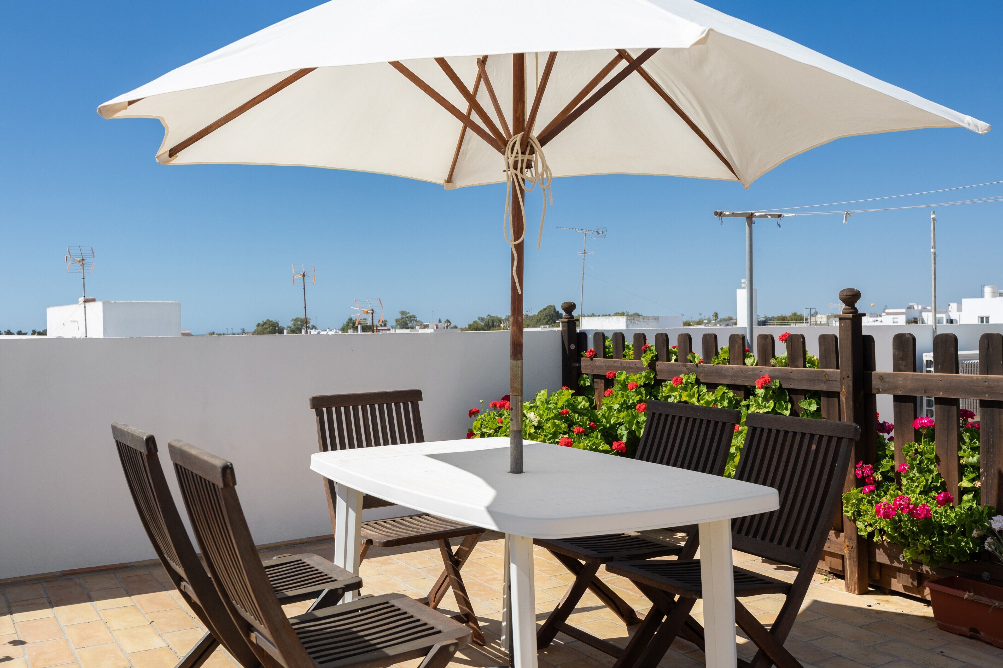 Un patio con una mesa blanca, cuatro sillas, una sombrilla y macetas con flores rojas.
