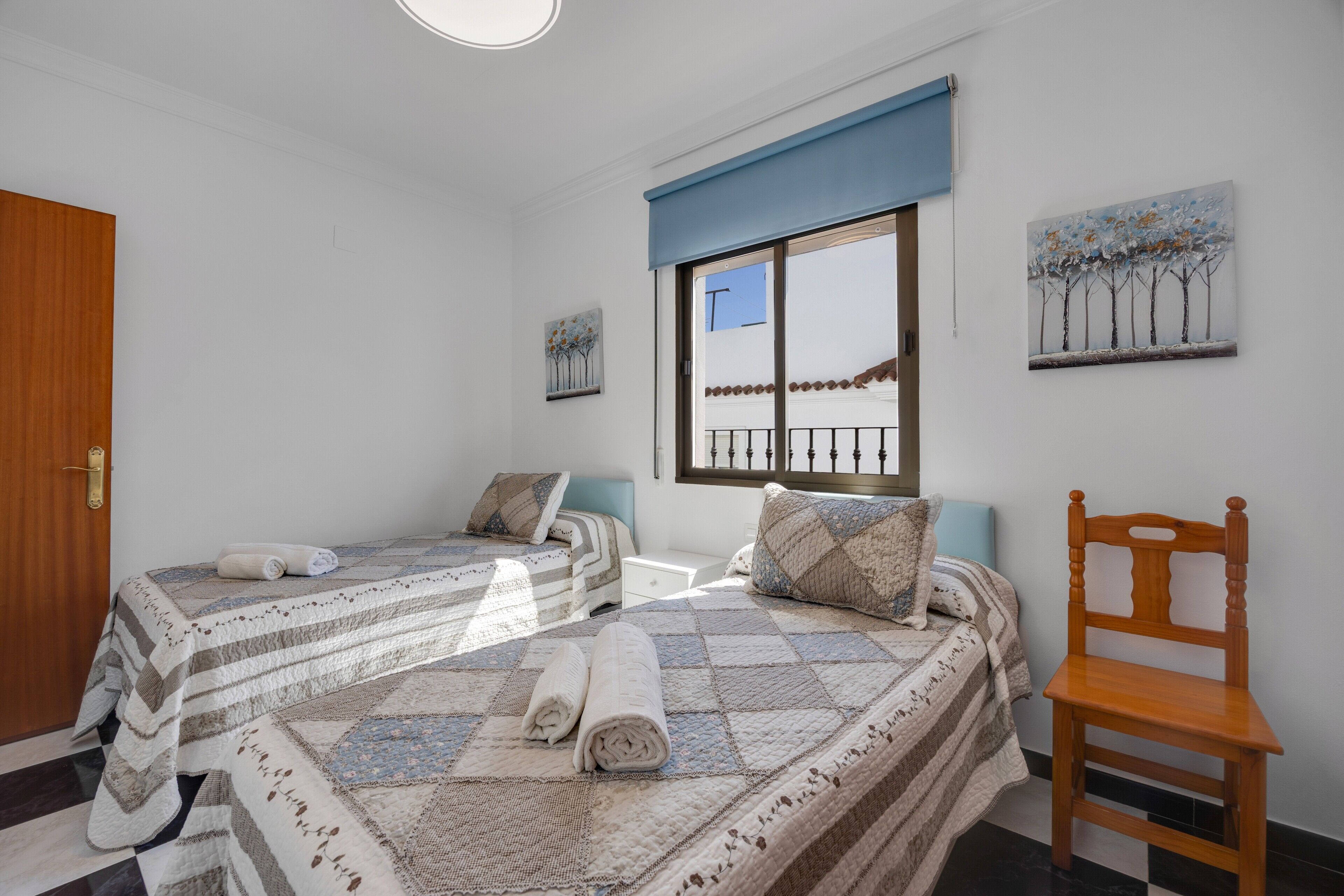 A bedroom with two beds, a wooden chair, a window with a view, and artwork on the walls.