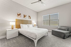 Une chambre à coucher avec un lit, des tables de chevet, une lampe, une fenêtre avec des stores et un canapé gris.