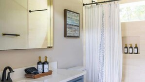 A bathroom with a white sink, blue cabinets, a black faucet, a white toilet, and a shower curtain. There are three bottles on the wall and a framed picture above the sink. - Countryside Escape | 5 minutes to Round Top | 2BR (Round Top)