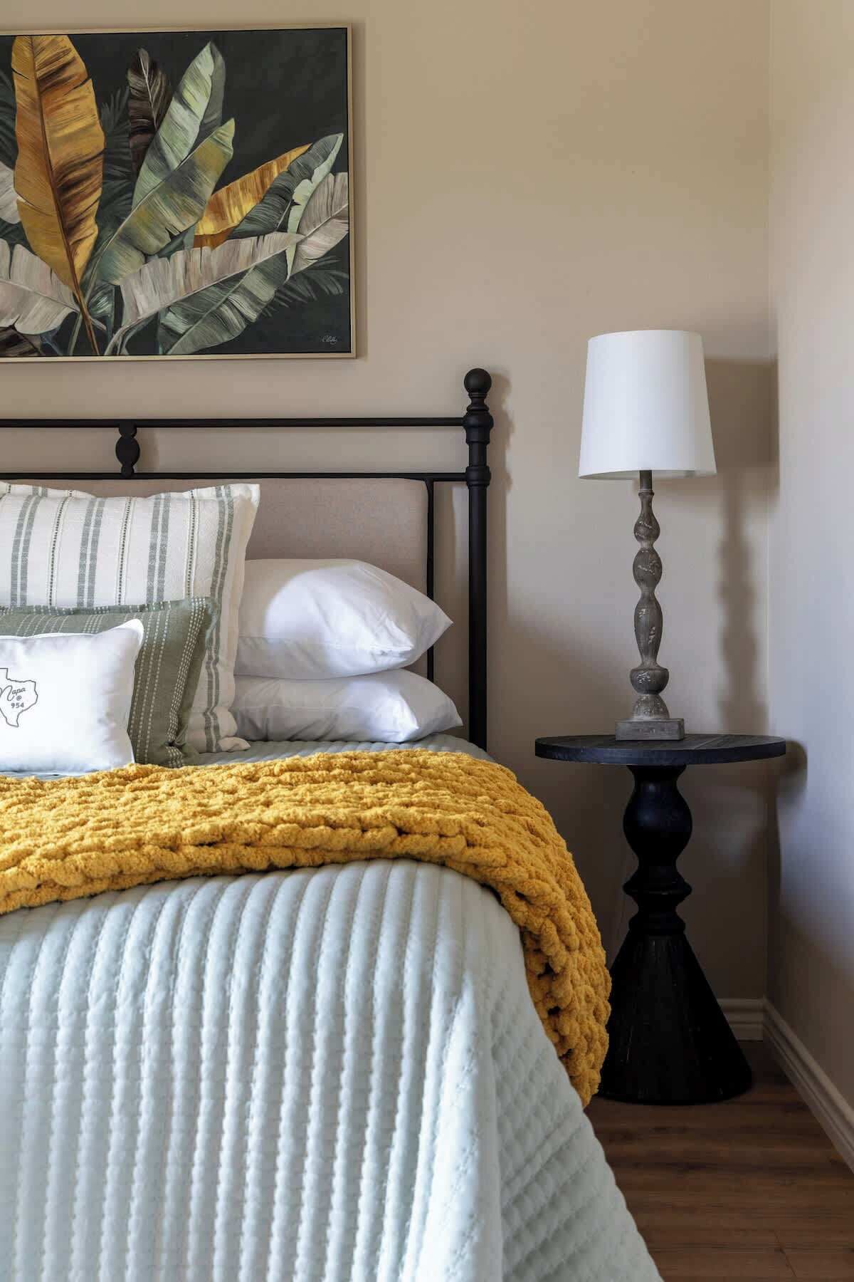 A neatly made bed with a yellow blanket, a bedside table with a lamp, and a framed botanical print on the wall.
