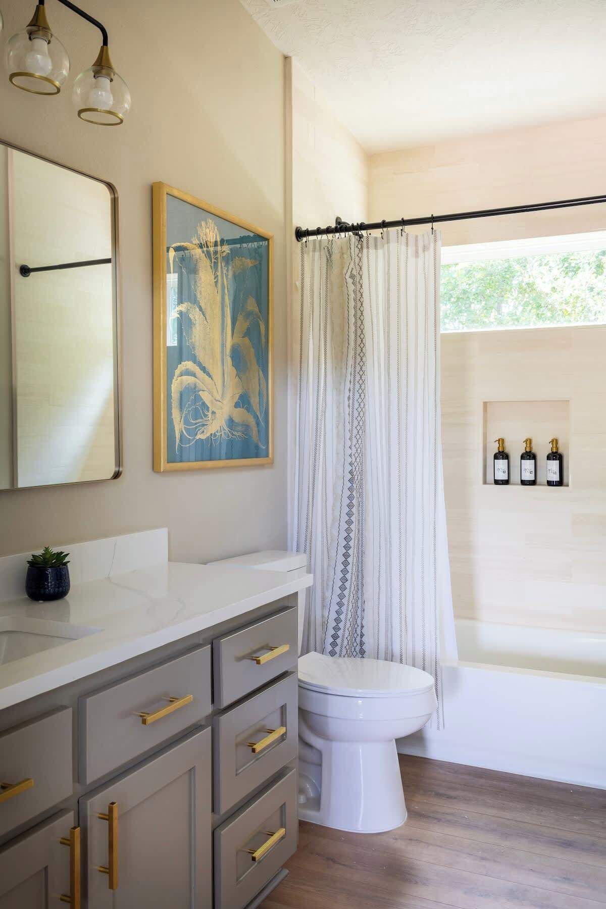 Une salle de bain avec un meuble-lavabo blanc, une œuvre d’art encadrée, un rideau de douche et des articles de toilette rangés sur une étagère.