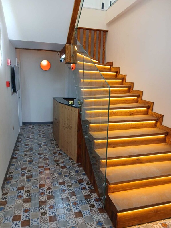 Una escalera con escalones de madera y barandillas de vidrio, que conduce a un rellano con techo plano y televisor montado en la pared.