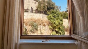 A window with curtains, a view of a tall tower, and a stone wall with vegetation. - The Little Prince Hotel (Antalya)