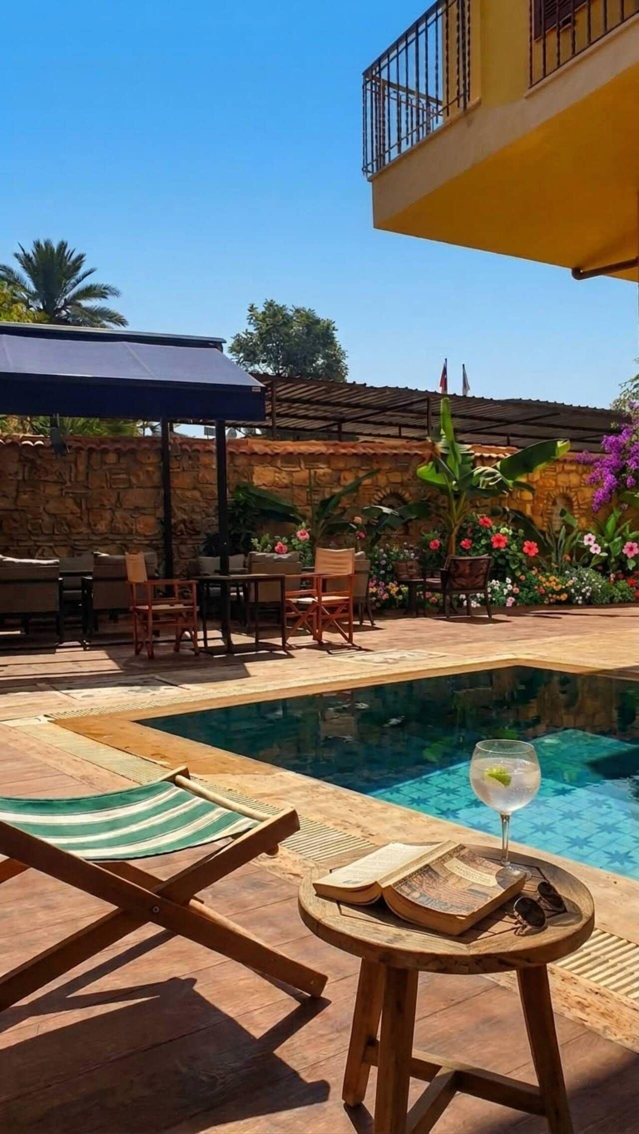 A pool area with lounge chairs, a table with a glass of water, and a stone wall with flowers.