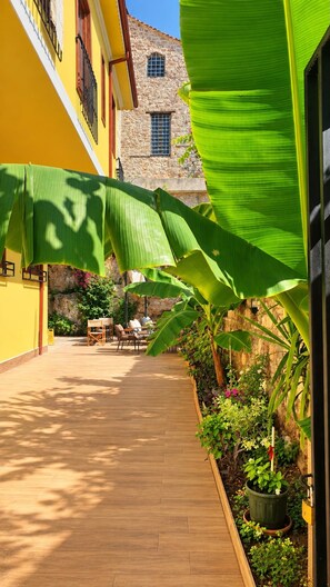 A wooden deck with a stone building in the background, surrounded by lush greenery and a yellow wall. - The Little Prince Hotel (Antalya)