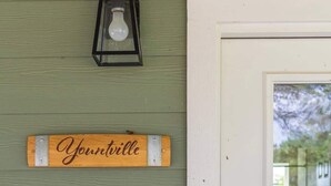 A wooden sign with the name "Yountville" mounted on a green wall, a black lantern above it, and a white door to the right. - Hidden Gem Near Round Top | 2BR Cottage (Round Top)