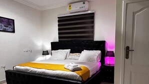 A neatly arranged bedroom with a black upholstered headboard, a bed with white linens and a yellow throw, a bedside table with a lamp, and a wall-mounted air conditioner.