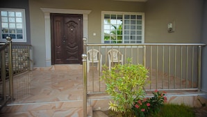 A front entrance with a marble floor, a dark wooden door, a white window with a grid pattern, and a small garden with pink flowers.