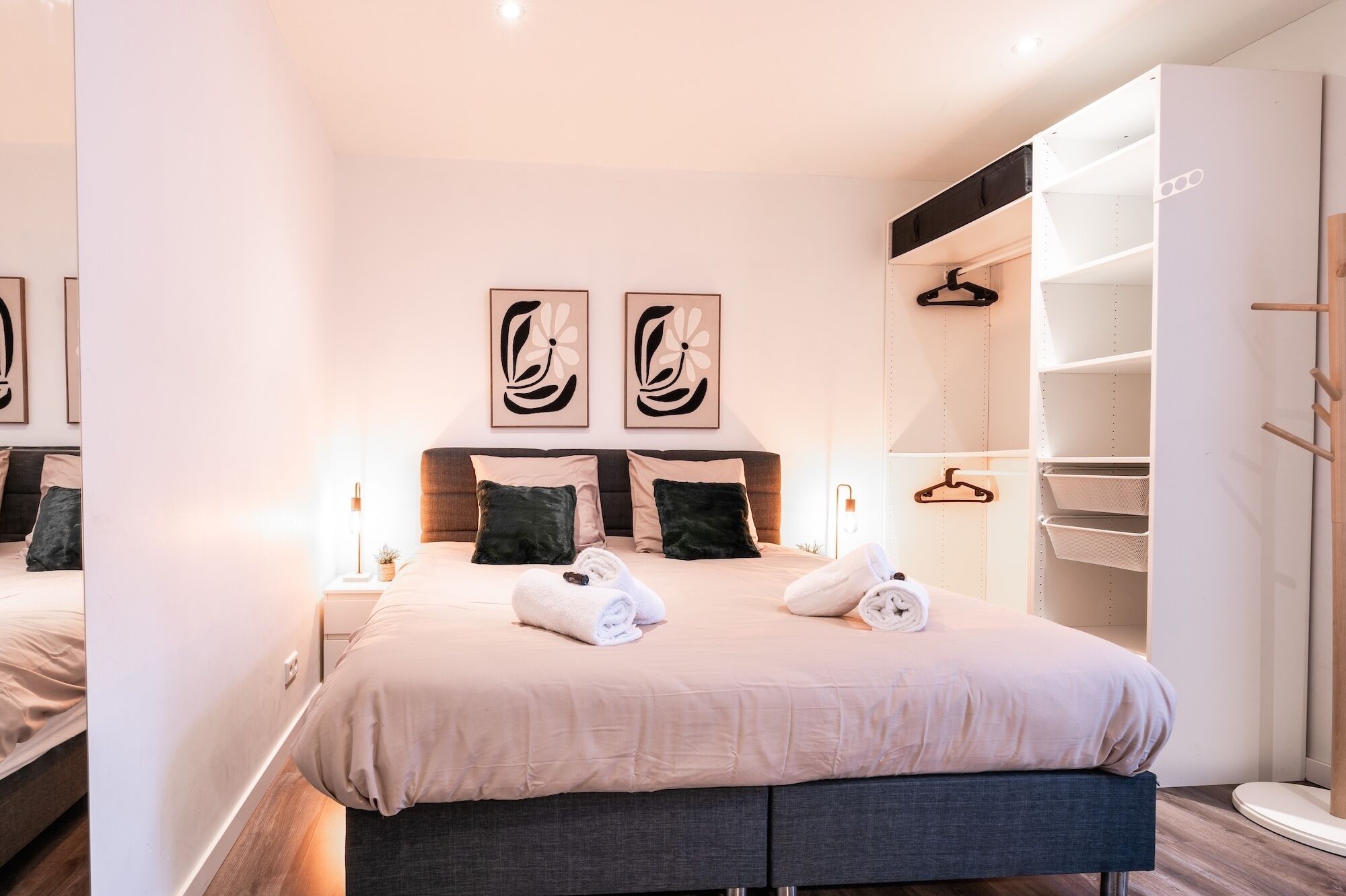 Un dormitorio moderno con una cama grande, dos obras de arte colgadas en la pared, una mesita de noche con lámpara y un armario empotrado con espacio para colgar ropa.