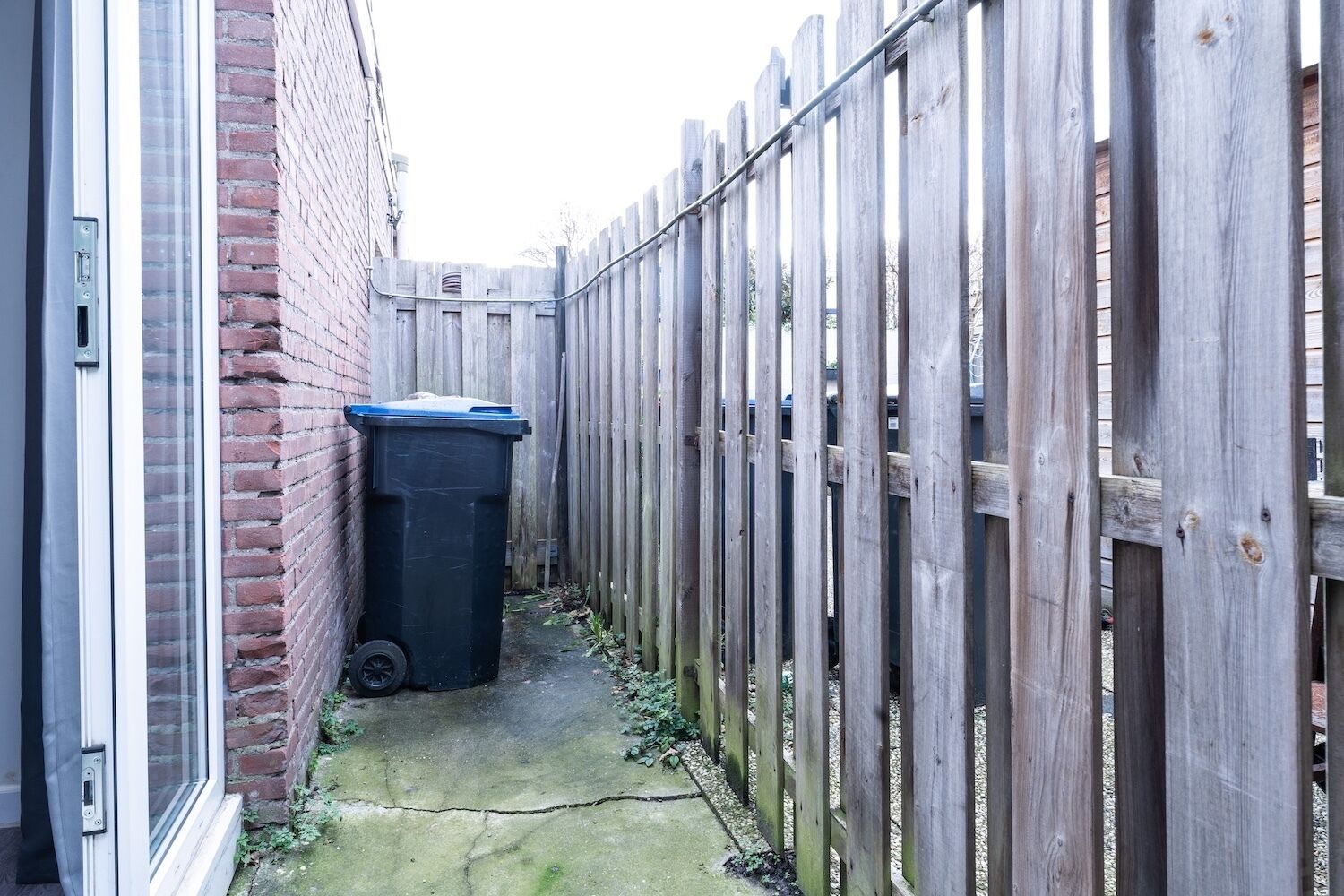 Un estrecho callejón con valla de madera, contenedor negro y pared de ladrillo.