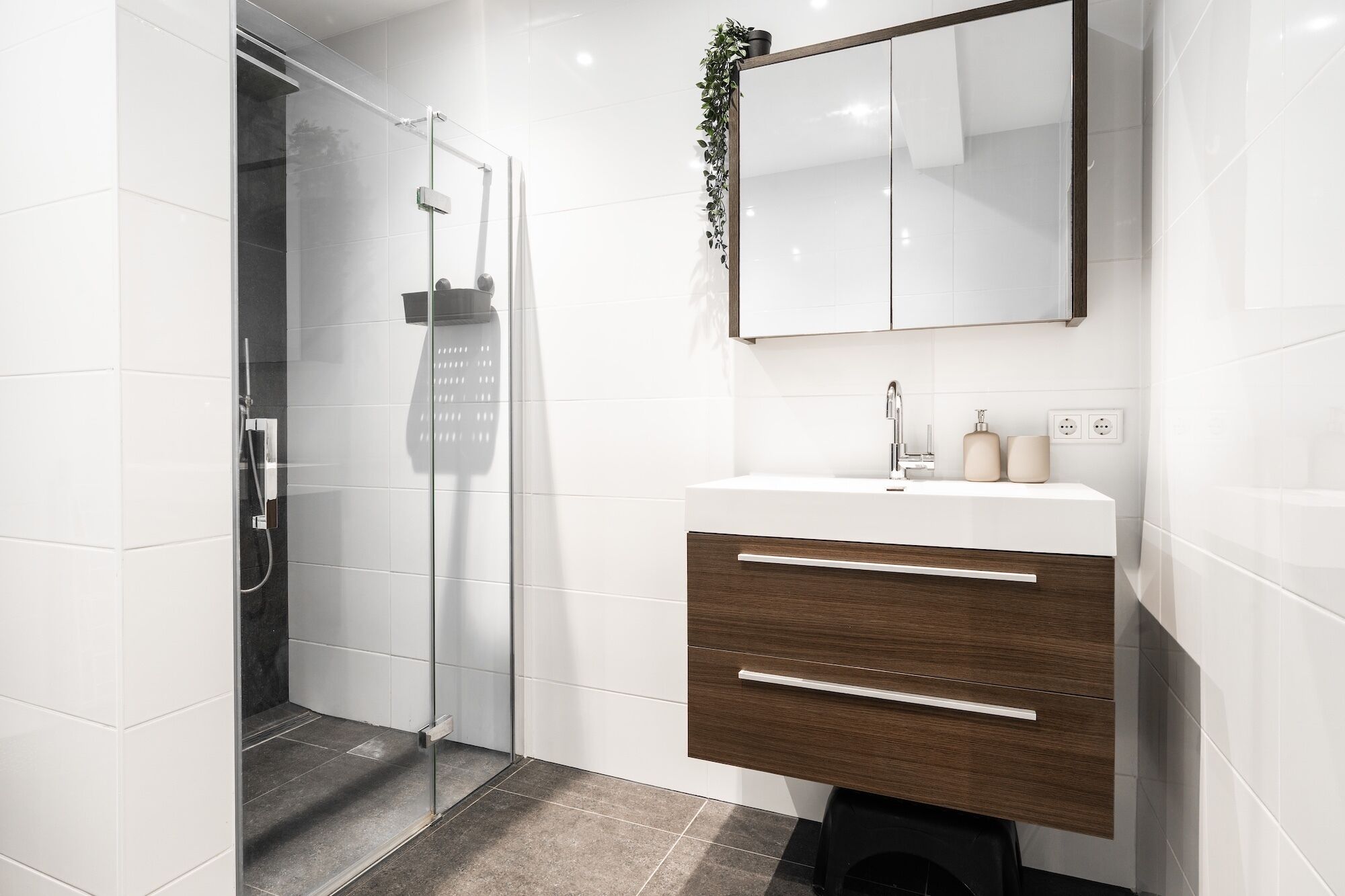 Un baño moderno con cabina de ducha de vidrio, tocador de madera con espejo y pared de azulejos blancos.