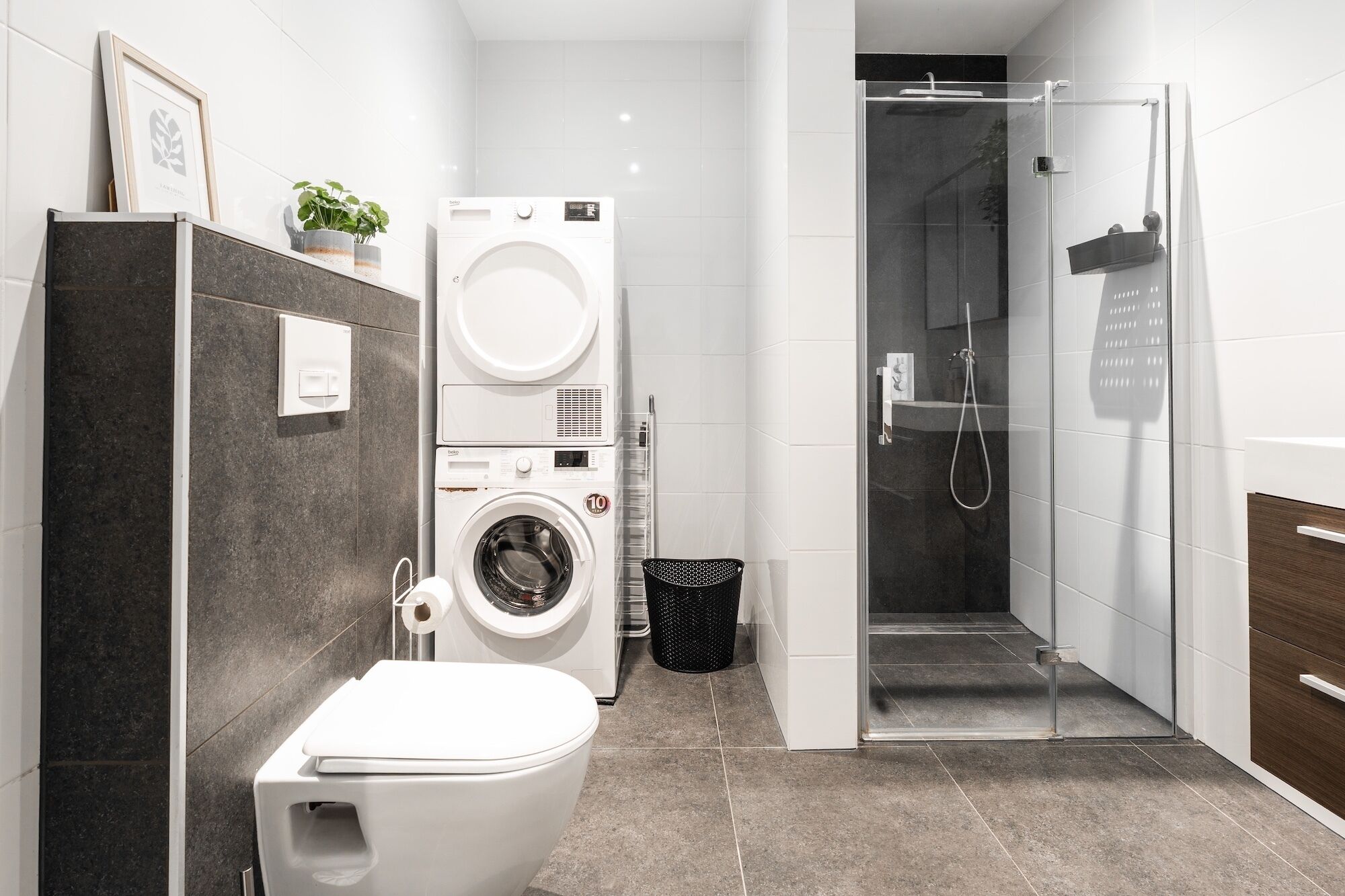 A compact bathroom with a toilet, stacked washing machine, and a glass-enclosed shower.